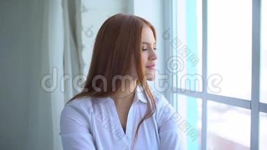 一个微笑的红头发女孩坐在家靠近窗户的肖像。 一个微笑的女学生的肖像。 女人微笑